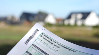 Ein Grundsteuerbescheid wird vor einer Wohnhaussiedlung hochgehalten.