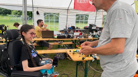 Luisa sitzt im Rollstuhl. Sie hat die Fernbediehung ihrer Drohne in der Hand. Ein Mann steht ihr gegenüber und stellt einen der Flugrotatoren ein.