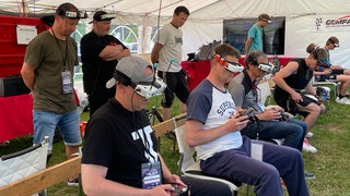 Teilnehmende Piloten sitzen mit einer Fernbedienung und einer VR-Brille auf einer Bierbank und steuern ihre Drohne durch den Parcour.