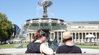 Eine Frau und ein Mann sitzen bei sommerlich-warmen Temperaturen auf einer Bank vor dem Springbrunnen auf dem Schlossplatz - da muss man doch glücklich sein?