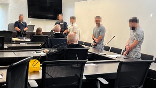 Landgericht Stuttgart: Angeklagte stehen in Handschellen im Gerichtssaal und werden dabei von Justizmitarbeitern bewacht.