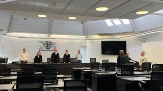 Im Gerichtssaal steht ein Angeklagter, der seine Partnerin getötet haben soll, und schaut zu den Richterinnen und Richtern.