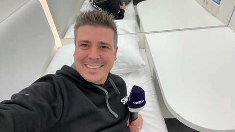SWR-Reporter Andy Dangel liegt beim Selbsttest der Schlafkabine mit Mikrofon in der Hand auf dem Bett 