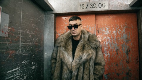 Der Rapper Haftbefehl steht mit einem Pelzmantel und einer Sonnenbrille in einem Fahrstuhl.