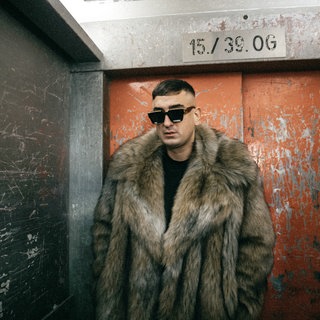 Der Rapper Haftbefehl steht mit einem Pelzmantel und einer Sonnenbrille in einem Fahrstuhl.