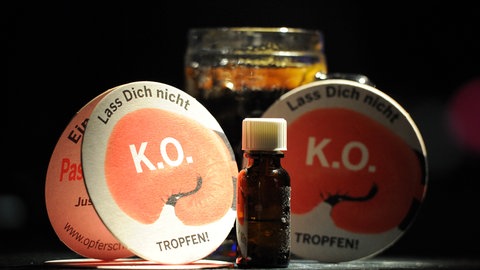 ein kleines Arznei-Fläschchen vor einem Cola-Glas mit Bierdeckeln einer Anti-K.o.-Tropfen-Kampagne (Symbolbild)