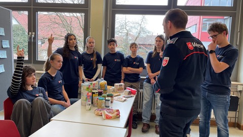 Neun Schülerinnen und Schüler des Georg-Büchner-Gymnasiums in Winnenden sitzen oder stehen um einen Tisch herum, auf dem ein Notvorrat zu sehen ist. In der Bevölkerungsschutz-AG lernen die Jugendlichen, wie man sich für den Notfall vorbereitet.