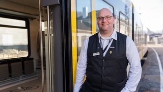 Ronald-Phillip Tolkiehn steht vor einem Zug von Arverio. Der Zugbegleiter ist von "Allianz pro Schiene" als "Eisenbahner des Jahres 2026" ausgezeichnet worden.
