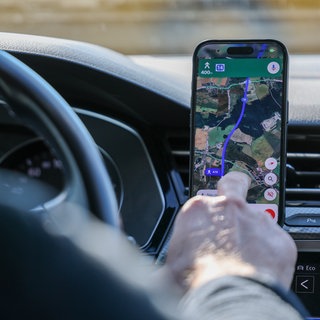 Mann tippt im Auto aufs Handy. Auf dem Handy ist eine Route von Google Maps.