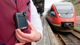 Ein Kundenbetreuer der Deutschen Bahn trägt eine Bodycam an der Dienstkleidung. Er steht neben einem Regionalzug. (Symbolbild)