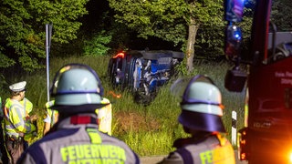 Das Unfallauto liegt auf der Seite an einem Baum, drum herum stehen Einsatzkräfte der Feuerwehr.