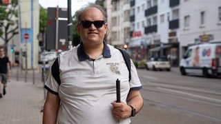 Georgios Karkalis steht auf dem Gehsteig und lächelt. Er trägt eine dunkle Sonnenbrille und hat seinen Blindenstock in der Hand.