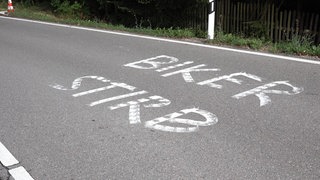 Auf einer Straße ist der Schriftzug "Biker stirb!" zu sehen.