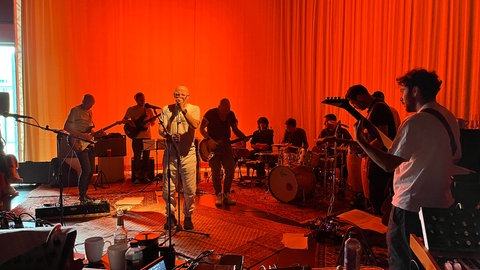 Studierende der Hochschule Offenburg proben gemeinsam mit Thomas D von den "Fantastischen Vier" vor einem rotangeleuchteten Vorhang.