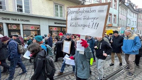 Eine Frau hält ein Plakat in die Höhe. Darauf steht: Domsingknaben - kostbares Kulturgut.