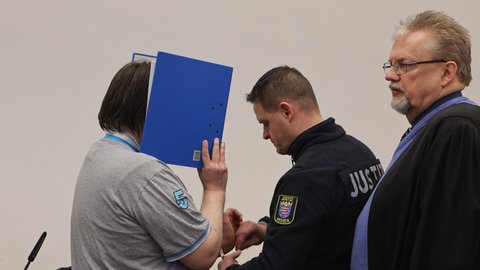 Der Angeklagte verbirgt sein Gesicht hinter einem Aktenordner und bekommt vor dem Prozess im Beisein seines Verteidigers Henner Maaß (r.) von einem Justizmitarbeiter die Handschellen abgenommen.