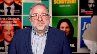 Mann in blauem Jackett steht vor einer Wand aus mehreren Wahlplakaten. Der Kommentator hat eine Brille und graue Haare. 
