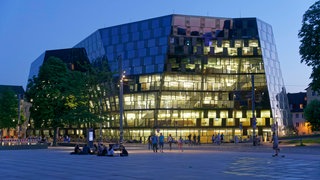 Das verspiegelte Gebäude der Unibibliothek Freiburg am Platz der alten Synagoge liegt beleuchtet in der Dämmerung.