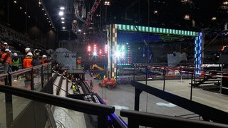 Eine Veranstaltungshalle mit Bühnenpodest. Auf einer Tribüne stehen Menschen mit Warnwesten und Schutzhelmen.