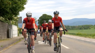 Zwei junge Frauen fahren auf ihren Rennrädern. Sie tragen Sonnenbrillen und Fahrradhelme. Hinter ihnen fahren weitere Menschen auf ihrem Rad.