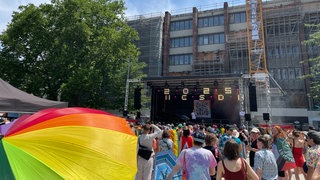 Im Vordergrund prankt ein Regenbpgen-Regenschirm. Der Christopher Street Day (CSD) starte im Freiburg mit einer Kundgebung auf dem Platz der Alten Synagoge. Im Hintergrund spricht Oberbrügermeister Martin Horn auf der Bühne.
