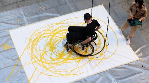 Ein kriegsversehrter Mann sitzt im Rollstuhl, malt dabei etwas Farbe auf ein großes darunter liegendes Blatt Papier.