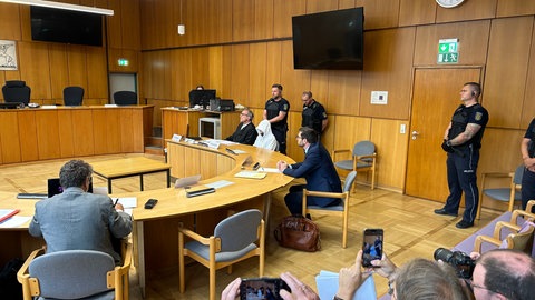 Das Bild zeigt einen Verhandlungssaal im Offenburger. Er sitzt neben seinem Anwalt, eine Kapuze tief ins Gesicht gezogen. Justizbeamte stehen am Rand des Gerichtssaals.