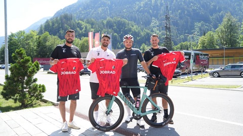 JobRad-Kurier Marius Frey liefert die neuen Trikots ins Trainingslager des SC Freiburg nach Schruns. M. Gulde, V. Grifo und Kapitän Ch. Günter nehmen die Trikots entgegen.  