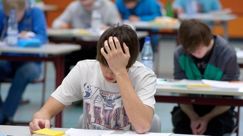 Im Vordergrund ist ein Schüler zu sehen, der an einem Tisch sitzt und offenbar mit allen anderen Schülerinnen und Schülern im Raum eine Prüfung schreibt.