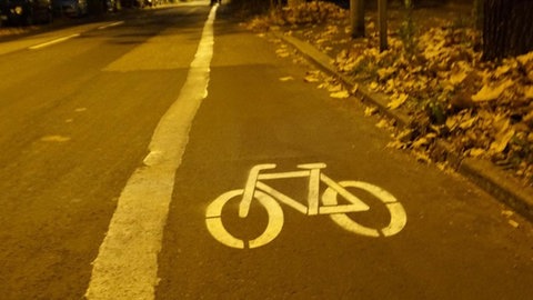 In weißer Farbe sind Begrenzungsteifen und ein Fahrrad auf die Straße gepinselt worden. Die Formen sind allerdings nicht ganz so akkurat wie gewöhnlich.