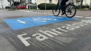 Auf eienr Straße ist das Logo für eine Fahrradstraße zu sehen. Ein weißes Fahrrad auf blauem Grund. Darüber fährt ein Fahrrad.