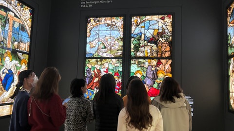 Eine Gruppe Menschen betrachtet ein buntes Kirchenfester aus dem Freiburger Münster.