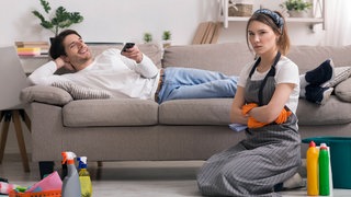 Ein Mann liegt auf dem Sofa und spielt auf seinem Handy während seine Partnerin putzt. 