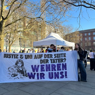 Zwei Demonstrierende halten ein Banner "Justiz & Uni auf der Seite der Täter? Wehren wir uns!"