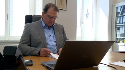 Ein Mann sitzt an einem Schreibtisch. Im Vordergrund ein aufgeklappter Laptop.