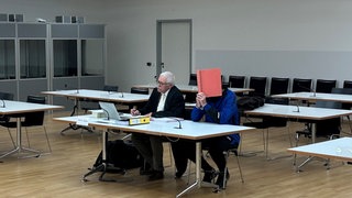 Der angeklagte Vater sitzt im Freiburger Landgericht neben seinem Verteidiger und versteckt sein Gesicht hinter einem Aktenordner.