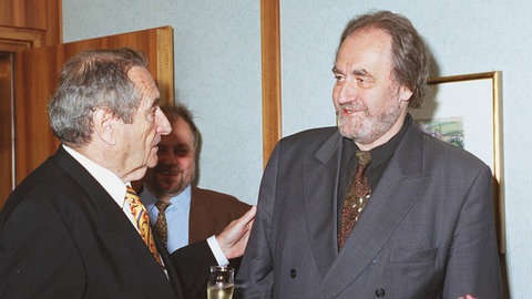 Walter Schultheiß mit einem Sektglas in der Hand, der sich mit Drehbuchautor Felix Huby - beide mit Schlips und Anzug - unterhält.