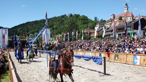 Eine blonde Frau in Rüstung auf einem fürs Turnier geschmückten Pferd vor der Kulisse der Stadt Horb, bei den Horber Ritterspielen 