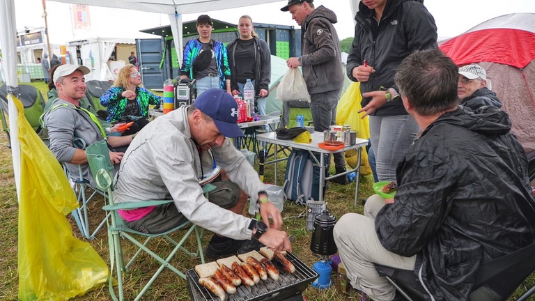 Würstchen-Grillen beim Southside-Festival 2024 in Neuhausen ob Eck. Viele Besucherinnen und Besucher haben sich gegen den Regen mit Gummistiefeln und Ponchos ausgestattet.