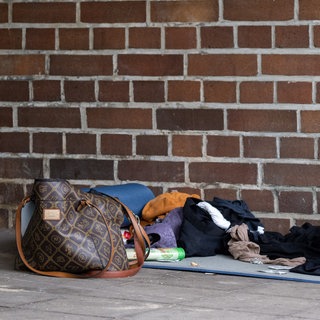 Verlassener Schlafplatz einer Obdachlosen. Eine Damenhandtasche, eine Isomatte und Kleidungsstücke liegen in einer Hofdurchfahrt.