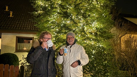 Zwei Männer vor einem Weihnachtsbaum. Die Besitzer Helmut und Willibald trinken Glühwein.