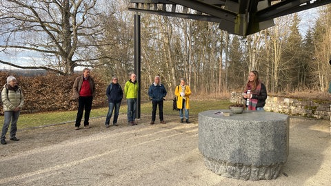 Lotta Brockel steht an einem steinernen Tisch vor der Gedenkstätte, im Halbkreis um sie einige Interessierte. 