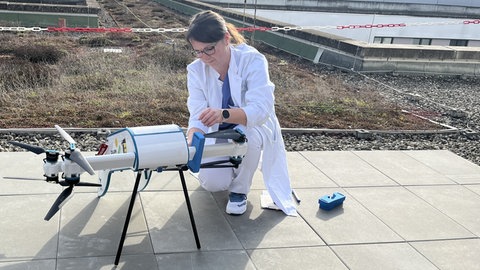 Eine Frau im weißen Kittel kniet neben der Labordrohne und sichert die Propeller ab.