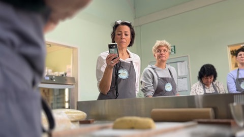 Zwei Teilnehmerinnen des Springerle-Backkurses in Münsingen stehen am Backtisch und haben sich ihre Schürzen angezogen. Eine von ihnen filmt mit der Handykamera die Kursleiterin.