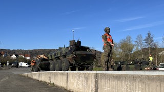 Ein Soldat in Uniform, vermummt und mit Warnweste steht vor einem Panzer. Er wartet, bis der Güterwagon frei wird.