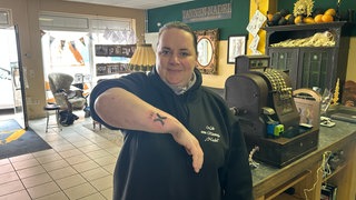 Eine Frau hält lächelnd ihren Unterarm in die Kamera. Darauf ist ihr neues Tattoo: "Tal X" - das Logo der Gartenschau 2025.