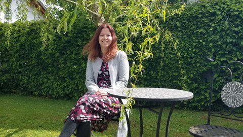 Vanessa Schmidt sitzt alleine auf einem Stuhl an einem Tisch im Garten. 