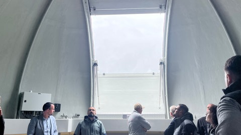 Menschen stehen in einer weißen Kuppel, im Hintergrund öffnet sich eine Luke. Beim Richtfest der Weltraumteleskope konnte man auch das Innere der Dachkuppel besichtigen.