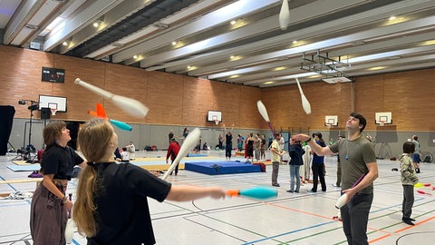 In einer Turnhalle verteilt jonglieren mehrere Gruppen mit Keulen und anderen Gegenständen. Sie haben sich zur Jonglier Convention getroffen