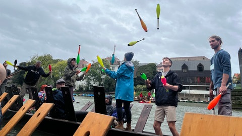Ein Stocherkahn auf dem Neckar, der Himmel grau. Teilweise mit Kaputzen jonglieren junge Menschen dennoch mit bunten Kegeln. Zweiter von rechts: Weltmeister (2023) Moritz Rosner.